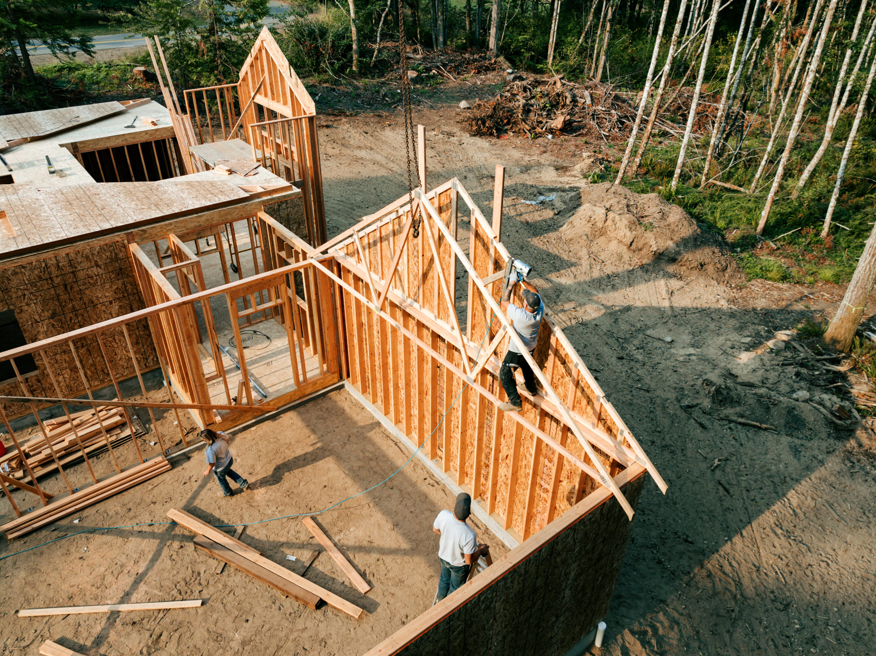 Workers build the frame of a new house project.  Bare plywood and beams as it is framed up from the foundation.  High lumber costs have affected the building process.  Shot in Washington state, USA.  High angle drone point of view.